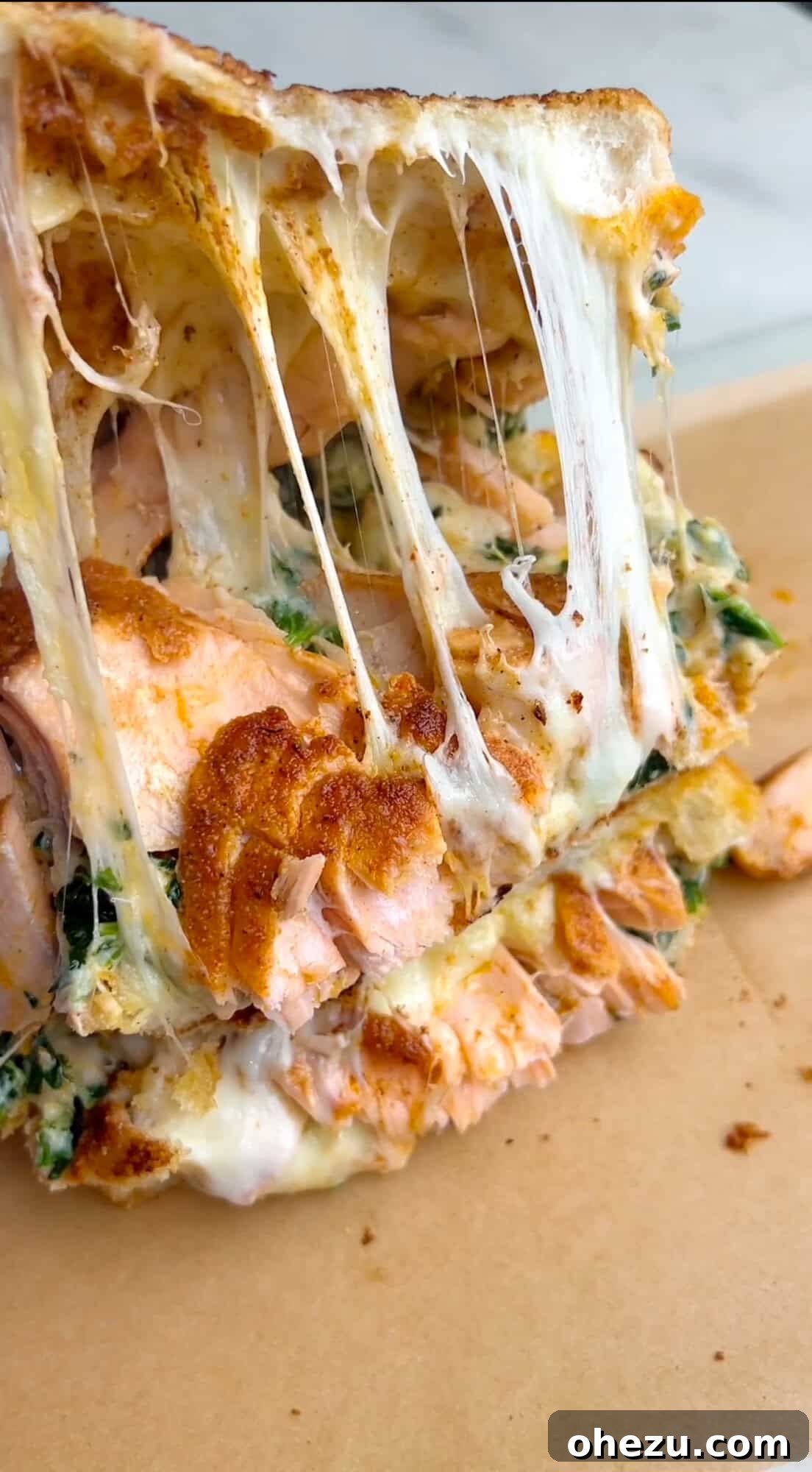 Lifting the top piece of bread from a salmon grilled cheese sandwich to reveal the perfectly melted cheese, flaky salmon, and creamy spinach artichoke spread inside.