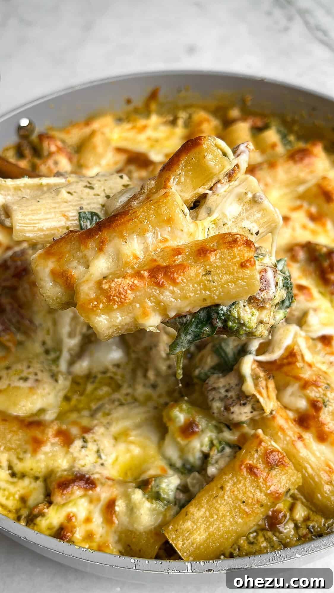 Close-up image of cheesy, golden brown rigatoni in creamy pesto sauce with chicken and fresh vegetables, served directly from the skillet.