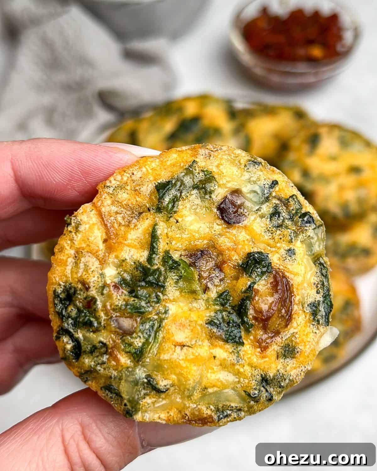 Enjoy a close-up of a freshly baked, perfectly fluffy kale and mushroom egg bite, ready for a delicious breakfast. A hand presenting a single, perfectly baked kale and mushroom egg bite to the camera.