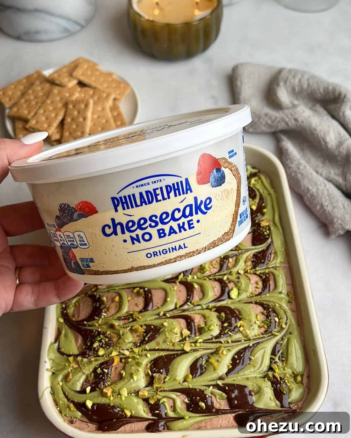 With layers of crispy kataifi, pistachio cream, and chocolate whipped cream, this Dubai chocolate cheesecake is a dream come true. A hand holding a slice of no-bake Philadelphia cheesecake, showcasing the beautiful layers.