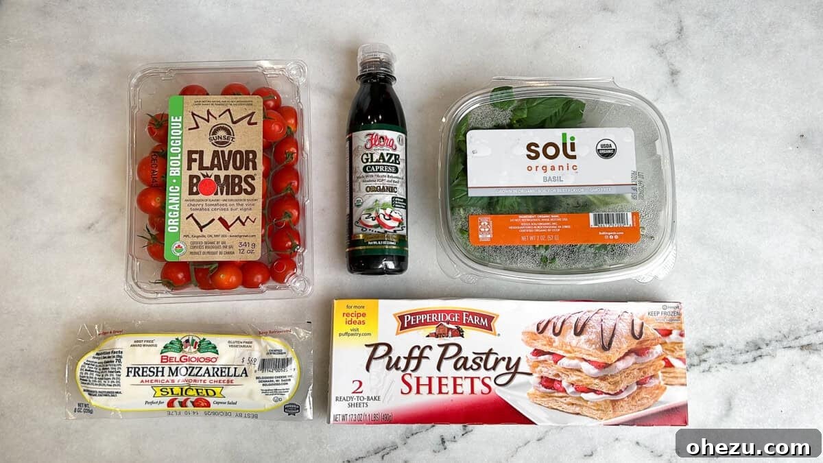 Overhead view of fresh ingredients for caprese bites: cherry tomatoes, balsamic glaze, basil, mozzarella cheese, and puff pastry laid out on a kitchen counter