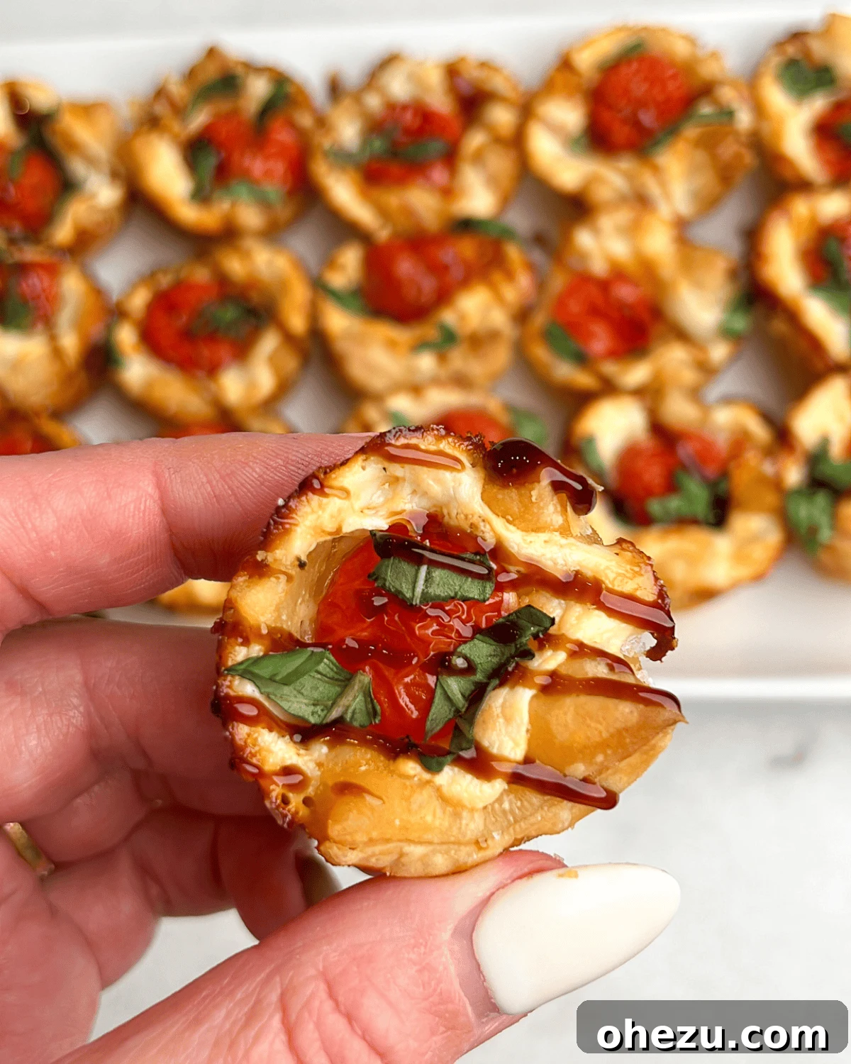 A hand reaching for and holding a single baked caprese bite, highlighting its perfect snackable size and appealing appearance.