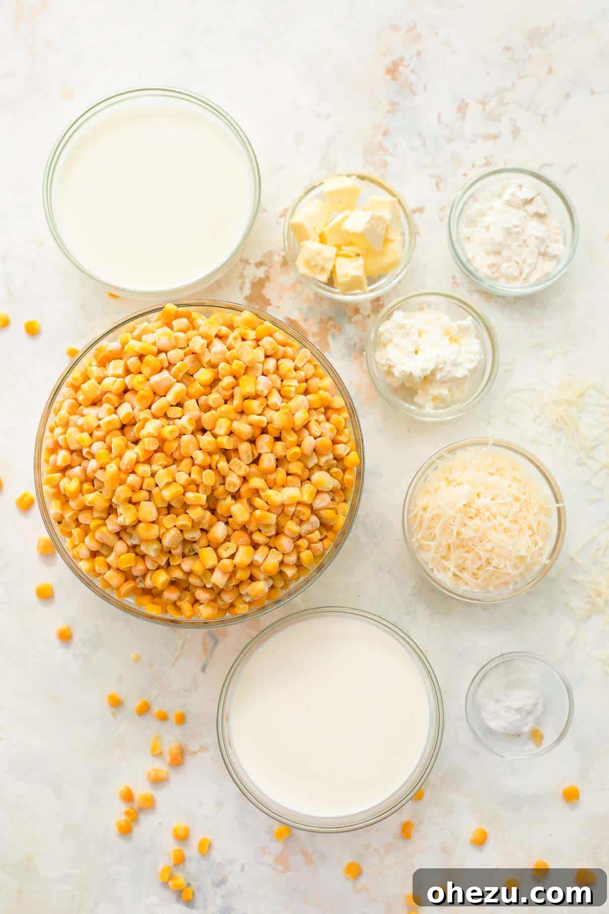 Glass bowls containing various ingredients for creamed corn: frozen corn, heavy cream, milk, butter, parmesan cheese, cream cheese, salt, pepper, and flour.