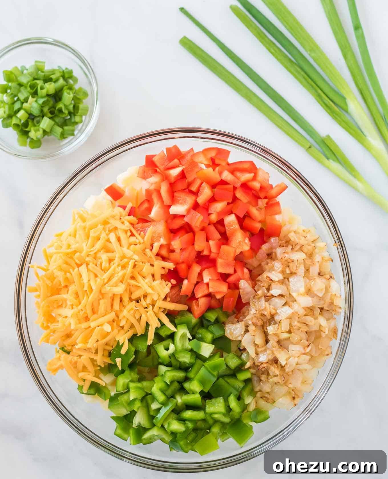 Sunrise Hash Brown Egg Bake 5 A large glass mixing bowl filled with a colorful mixture of thawed hash browns, diced bell peppers, sautéed onions and garlic, and half of the grated cheddar cheese, ready to be combined.