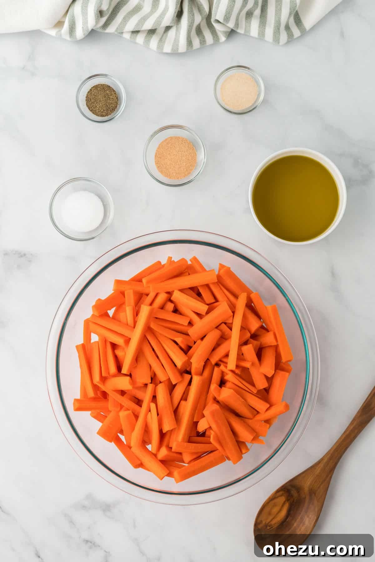 The Ultimate Roasted Carrots 5 Precisely cut carrots in a large mixing bowl, accompanied by small individual bowls of olive oil and various spices (garlic powder, onion powder, salt, pepper) awaiting combination.