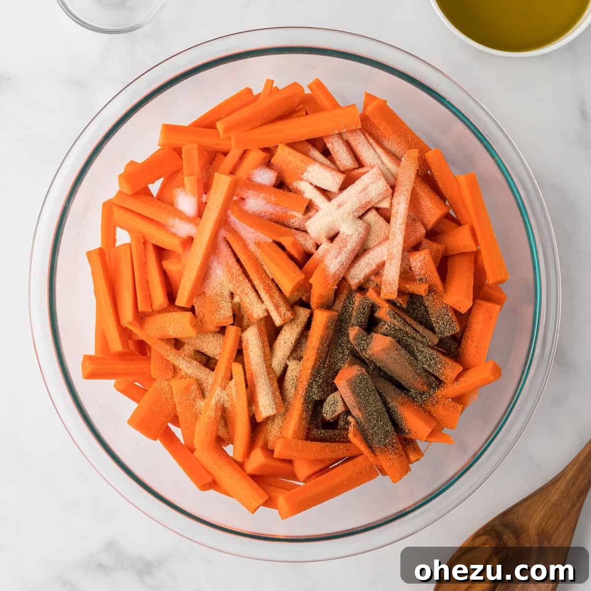 The Ultimate Roasted Carrots 6 A hand sprinkling a blend of spices, including garlic powder, onion powder, salt, and pepper, over uniformly sliced carrots in a mixing bowl.