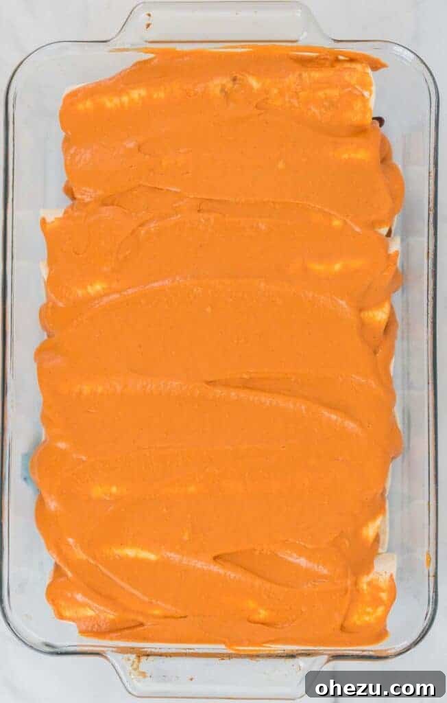 Rolled vegan enchiladas neatly arranged in a greased casserole dish, generously topped with the creamy cashew queso sauce, ready for baking.