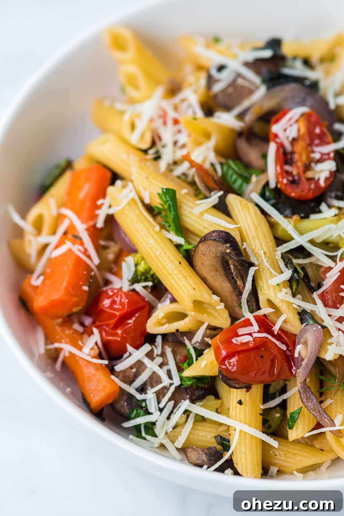 A white bowl filled with roasted vegetable pasta, garnished generously with freshly grated parmesan cheese.