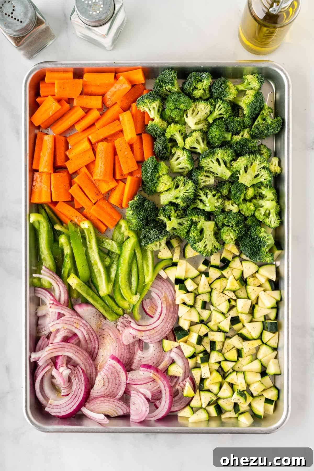 Chopped carrots, broccoli, bell peppers, onion, and zucchini spread on a baking sheet, seasoned with olive oil, salt, and pepper.