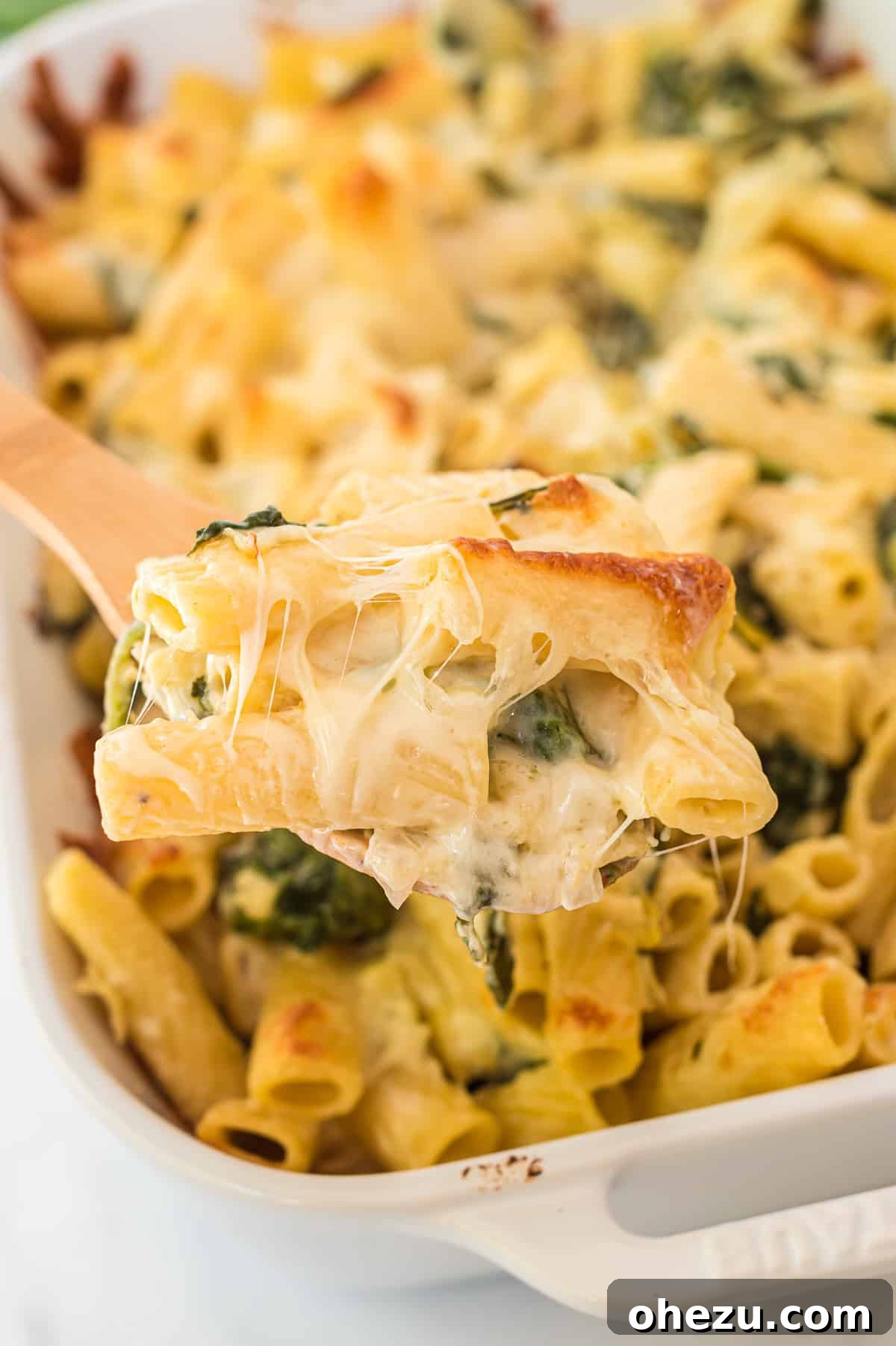 Creamy Spinach Artichoke Pasta Casserole 2 A wooden spoon scooping creamy spinach artichoke pasta bake from a white baking dish, showing melted cheese and pasta