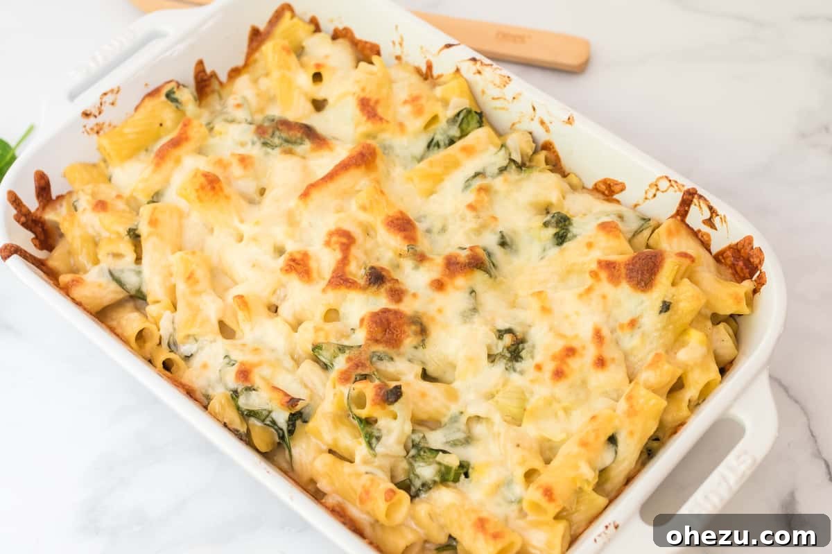 Creamy Spinach Artichoke Pasta Casserole 3 Close-up of a white baking dish filled with golden-brown, bubbly spinach artichoke pasta bake