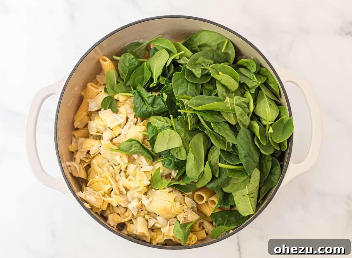 Creamy Spinach Artichoke Pasta Casserole 5 White Dutch oven with rigatoni pasta, topped with chopped artichoke hearts and fresh spinach, waiting to be mixed into the sauce