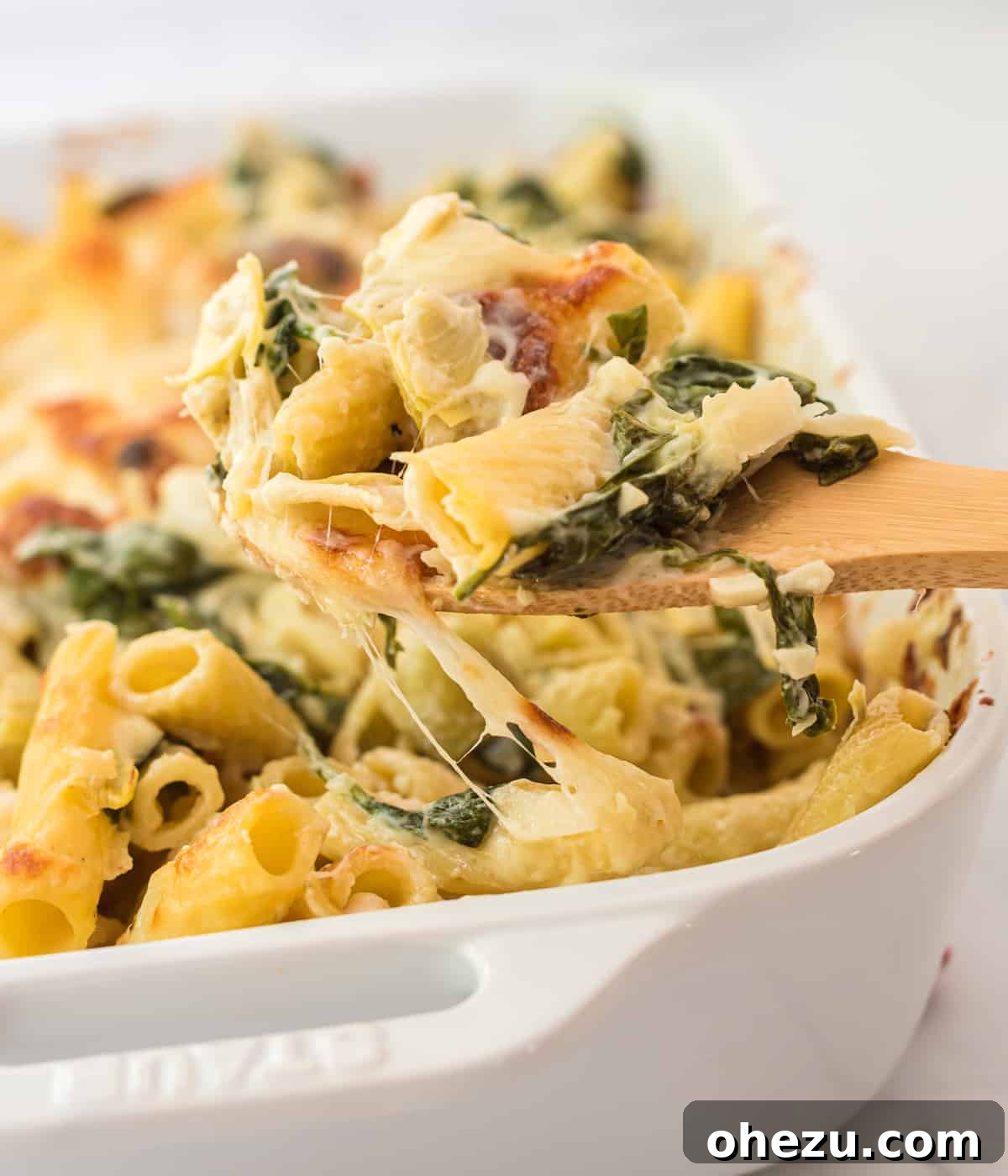 Creamy Spinach Artichoke Pasta Casserole 7 A wooden spoon taking a generous scoop of spinach artichoke pasta bake from a baking dish, highlighting the creamy texture and melted cheese