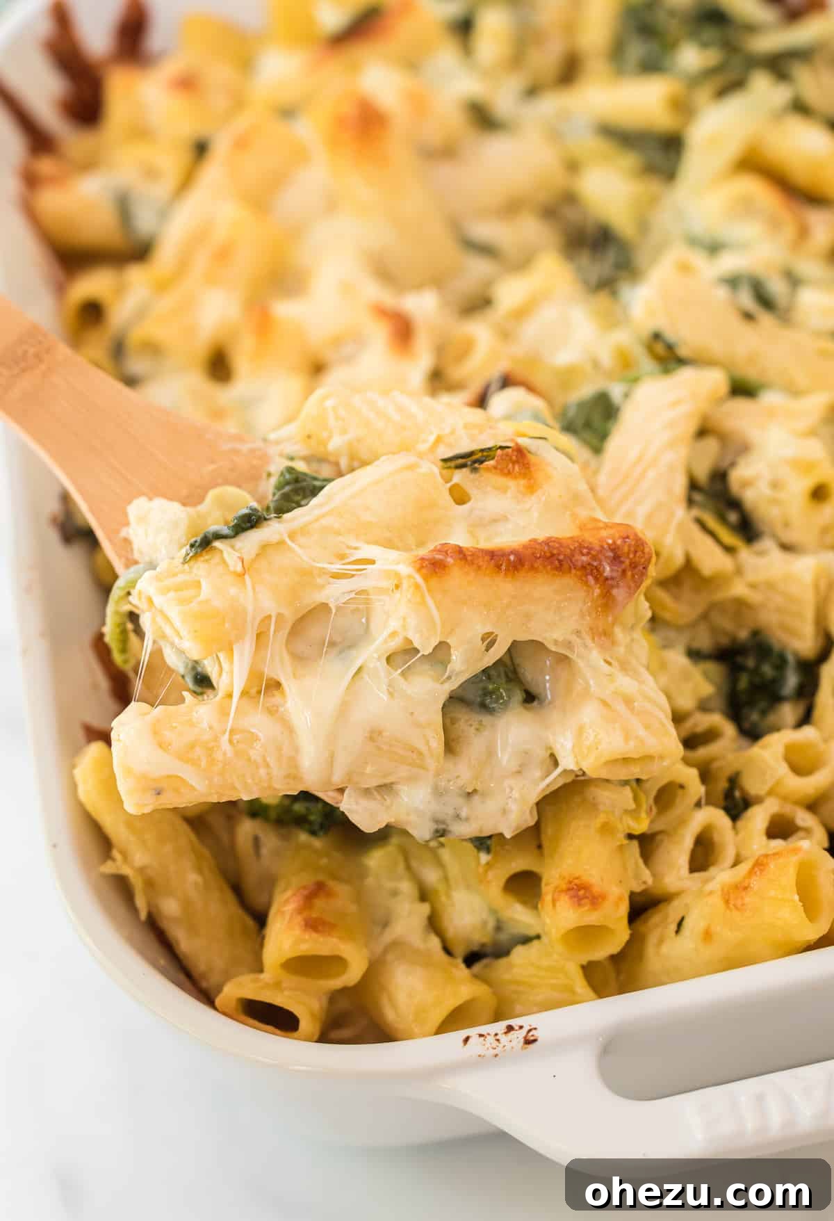 Creamy Spinach Artichoke Pasta Casserole 8 A second image of a wooden spoon taking a scoop of spinach artichoke pasta bake, emphasizing the texture and warmth