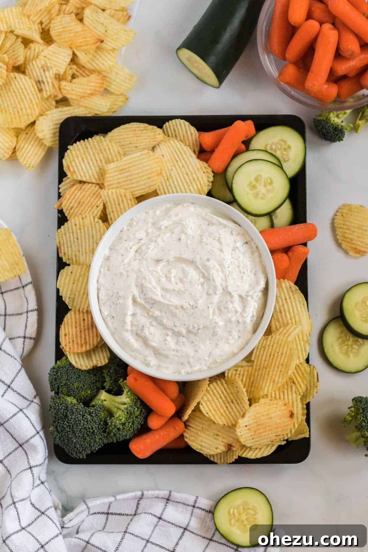 A generous bowl of creamy chip dip surrounded by a colorful array of dippers including ruffle chips, broccoli florets, baby carrots, and cucumber slices, all arranged appealingly.