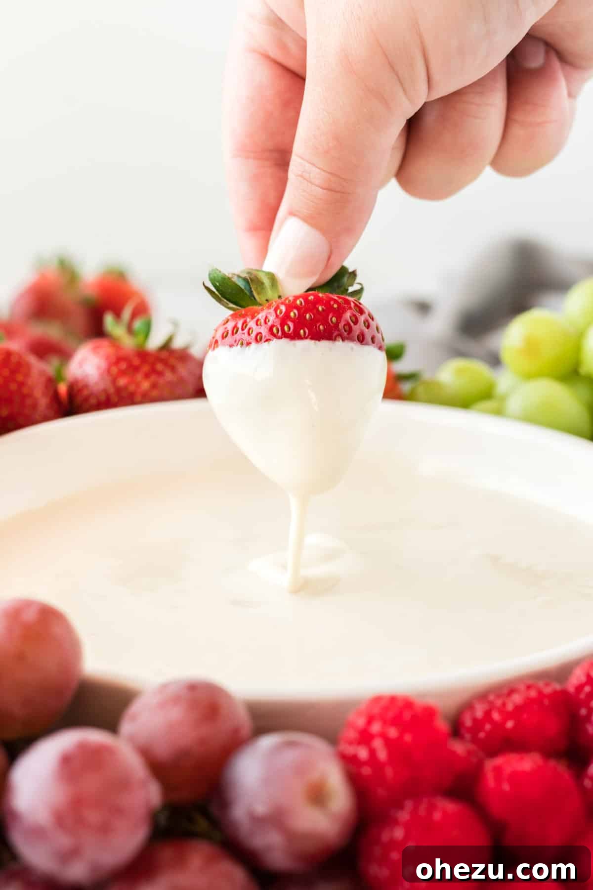 A hand gently dipping a fresh strawberry into a bowl of rich, smooth cream cheese yogurt dip.