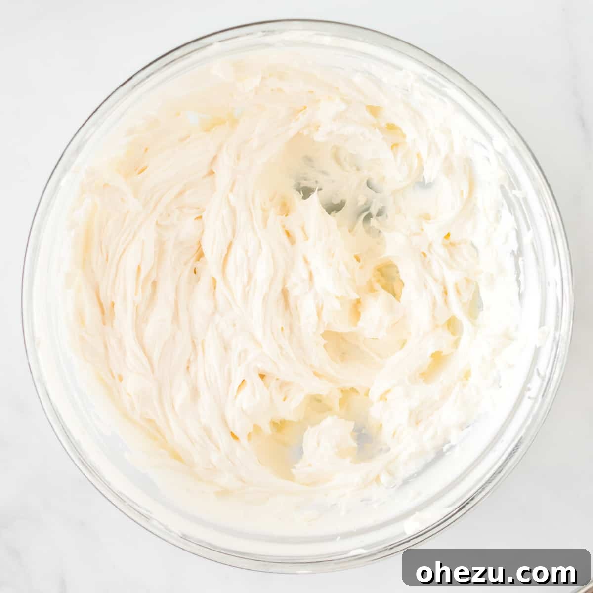 Softened cream cheese being beaten until fluffy in a large mixing bowl with an electric mixer.