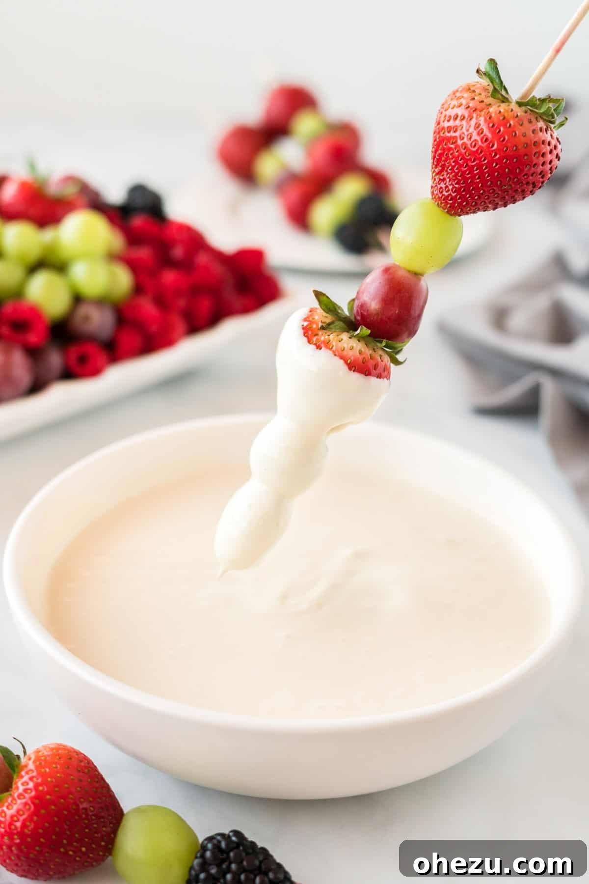 A colorful fruit skewer with strawberries, grapes, and blackberries being dipped into a bowl of creamy fruit dip.
