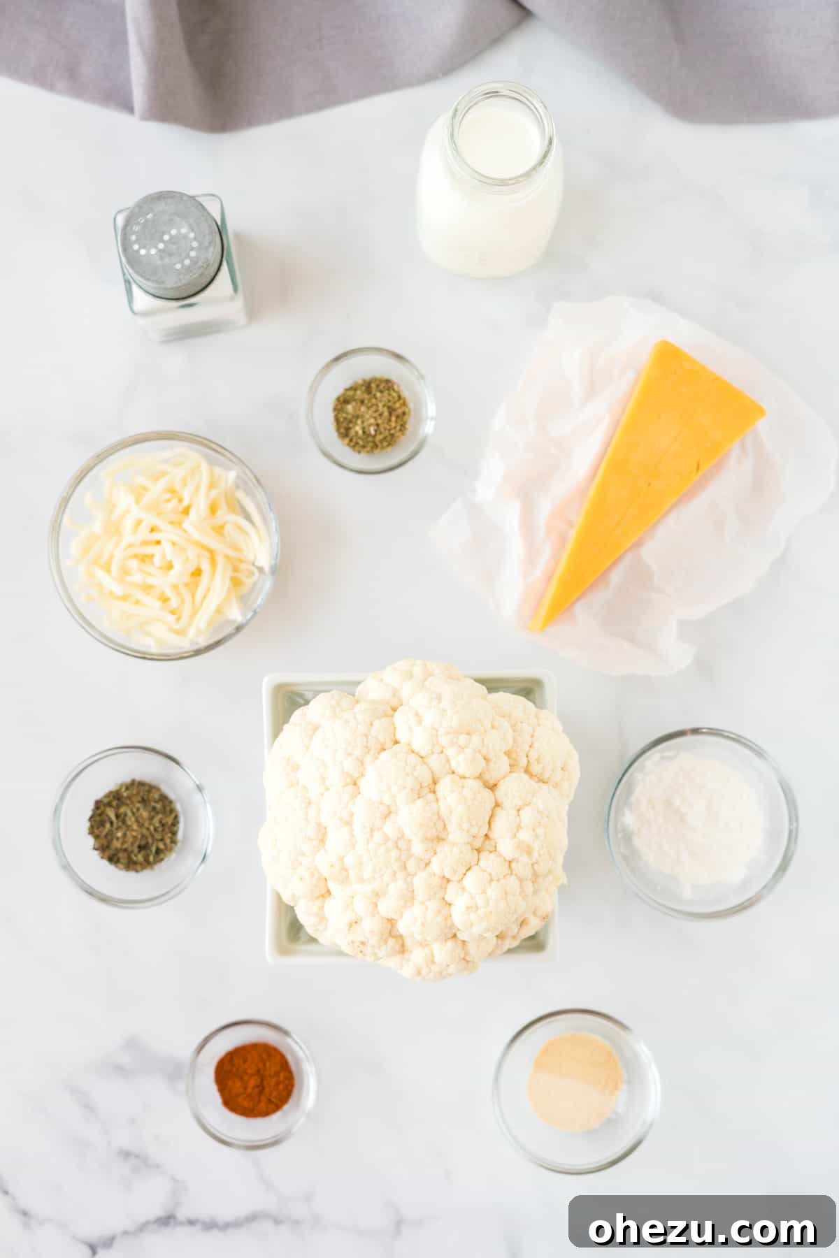A selection of fresh ingredients laid out on a table, including a head of cauliflower, various cheeses, milk, and an array of spices, ready for making cheesy cauliflower.