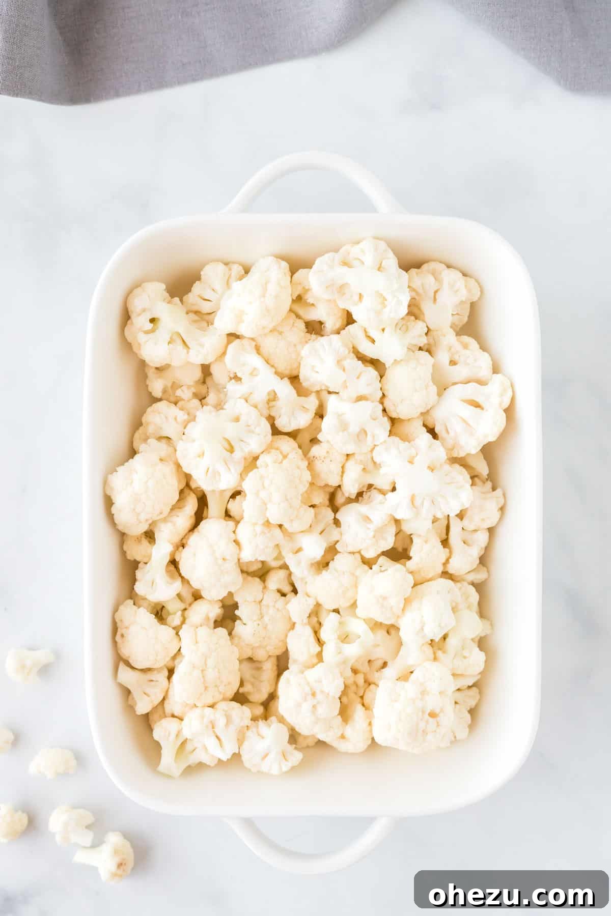 Freshly chopped cauliflower florets arranged in a white baking dish, lightly coated with olive oil spray, ready for roasting.