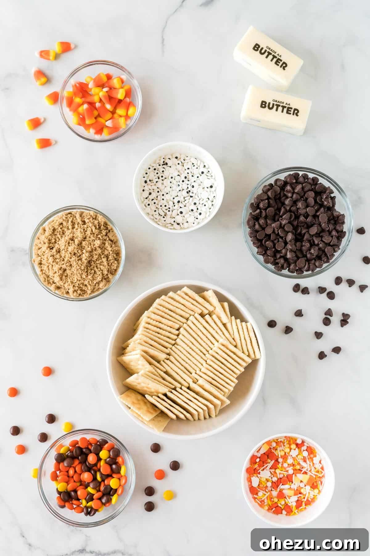 A curated selection of ingredients laid out on a clean surface, including saltine crackers, butter, brown sugar, chocolate chips, and various Halloween candies like eye candies, candy corn, and sprinkles.