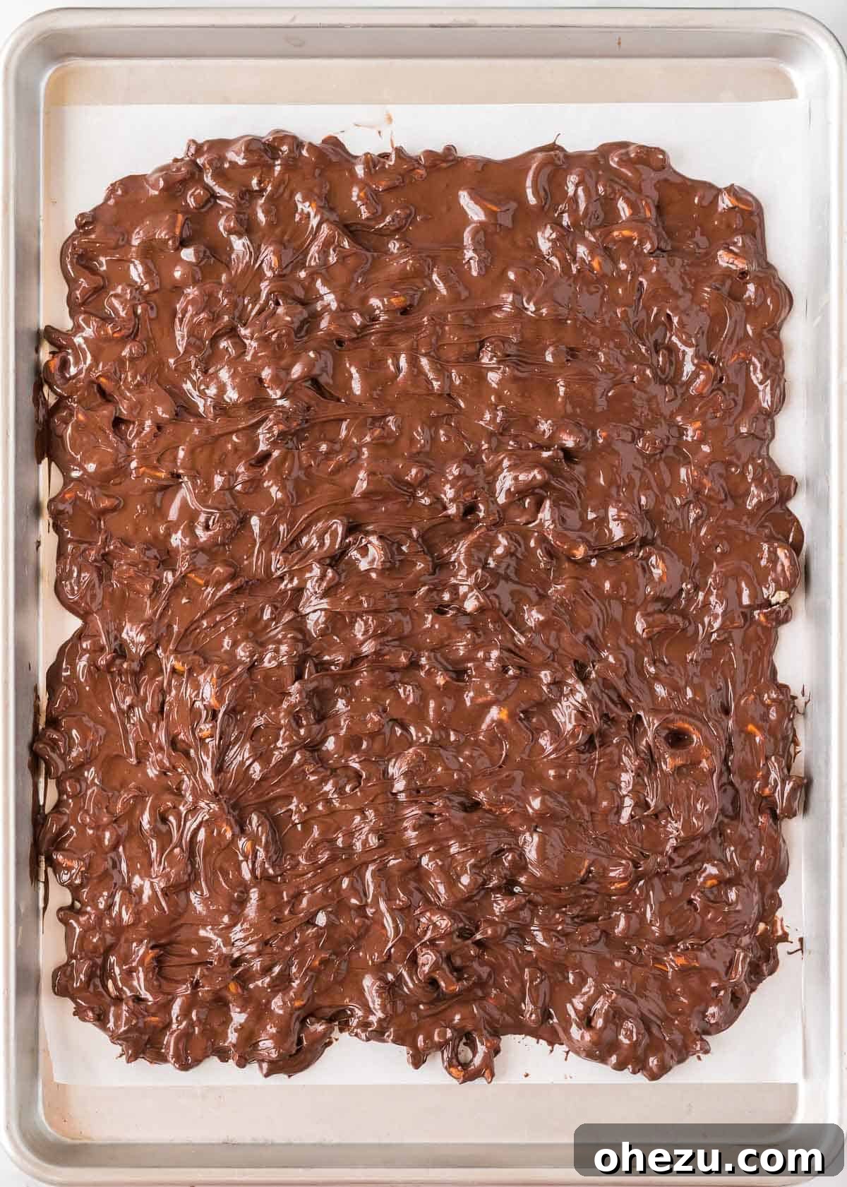 Melted chocolate and pretzels spread out on a baking sheet lined with parchment paper