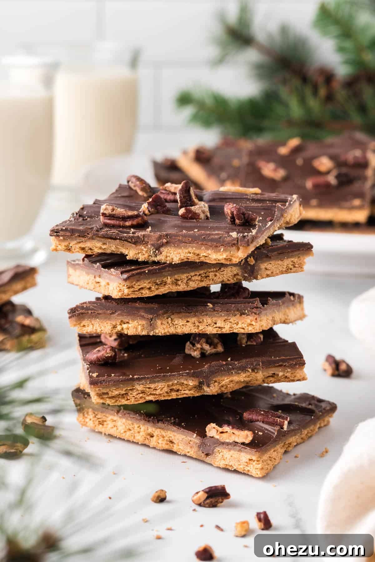 Stacked pieces of golden graham cracker toffee candy drizzled with rich chocolate and topped with toasted pecans, highlighting its irresistible layers.