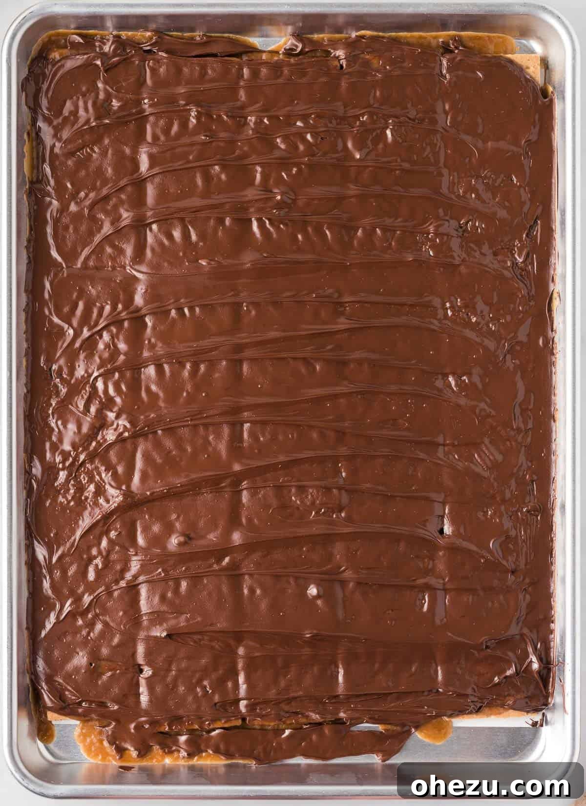 A rubber spatula expertly smoothing out the recently melted chocolate, creating a uniform, glistening, and inviting surface over the toffee layer.