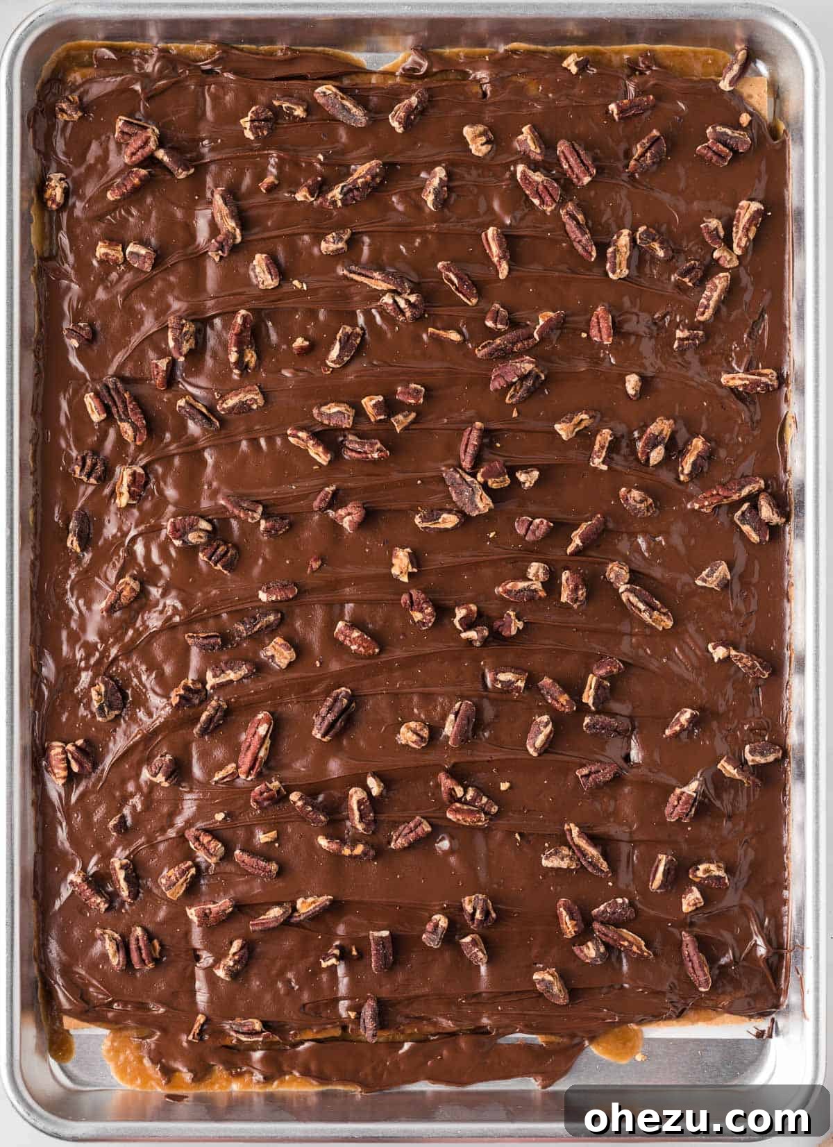 Toasted pecan pieces being generously sprinkled over the still-warm, freshly melted chocolate layer of the toffee, adding a delightful crunch and rustic appeal.