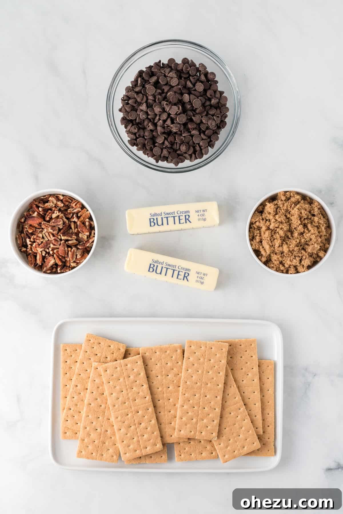 A flat lay photograph displaying all the simple, high-quality ingredients laid out on a surface: graham crackers, sticks of butter, a bowl of brown sugar, a bowl of chocolate chips, and a bowl of pecan pieces.
