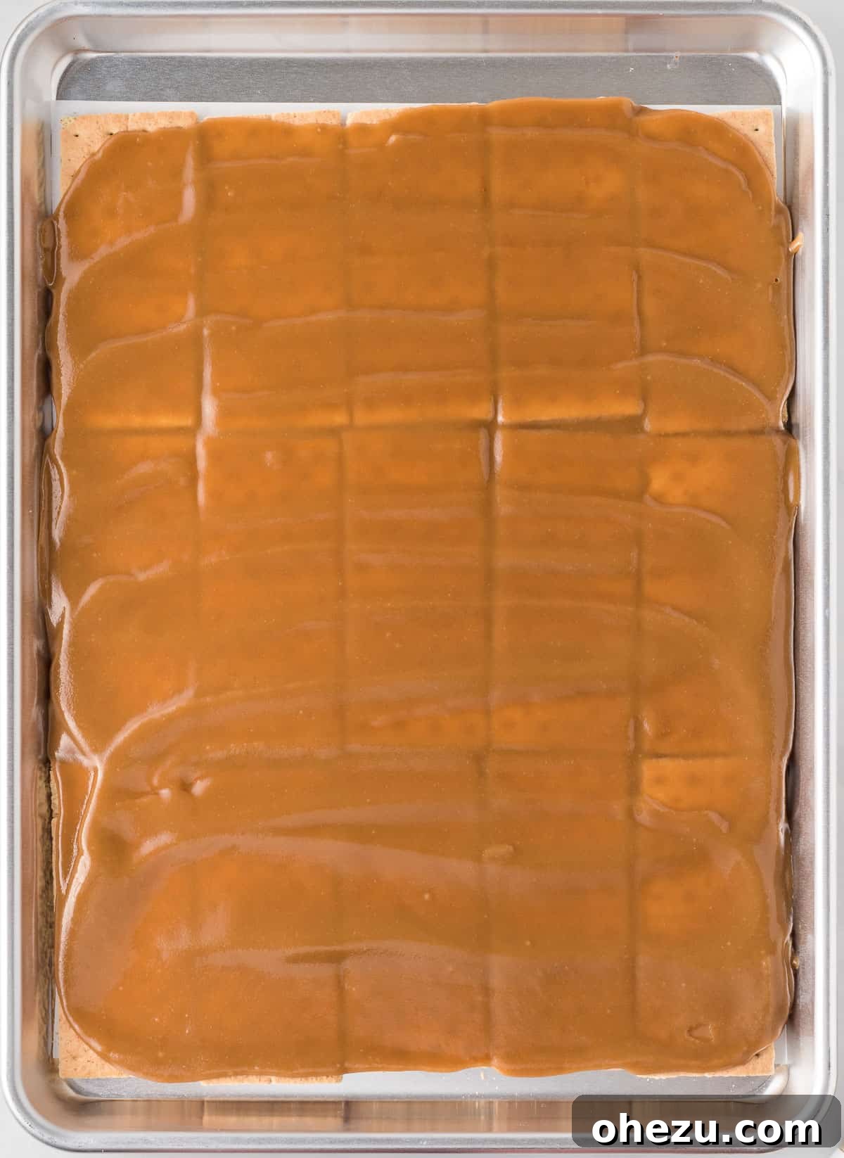 A rubber spatula meticulously spreading the hot, glistening, golden toffee mixture evenly and smoothly over the arranged graham crackers on the baking sheet.