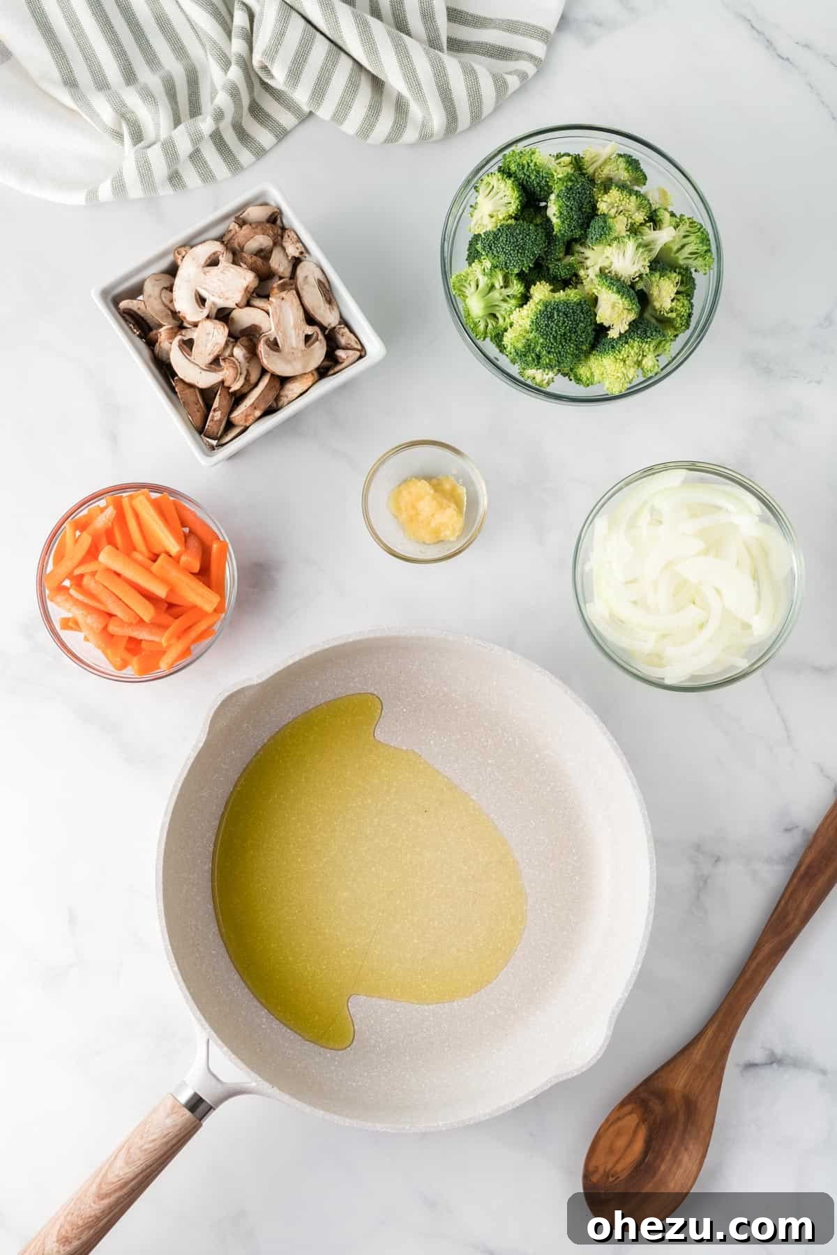 A large skillet with olive oil heating over medium-high heat, surrounded by organized bowls of prepped yellow onion, carrots, broccoli, garlic, and sliced mushrooms, ready to be added for stir-frying.