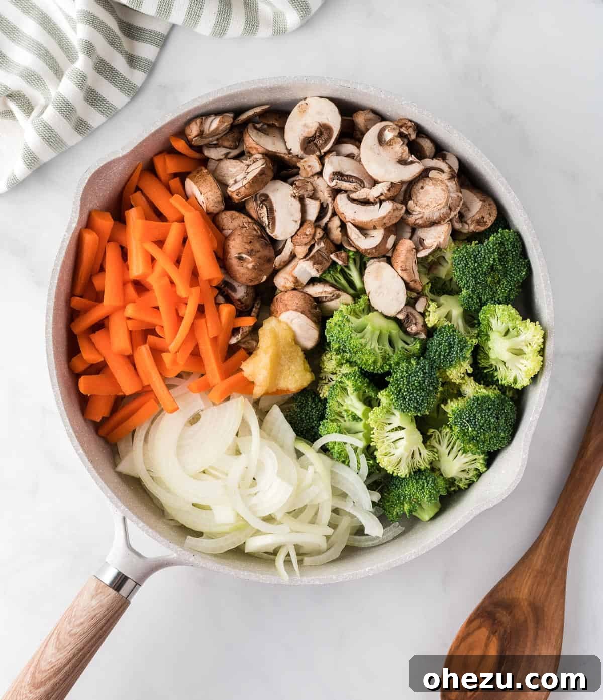 A large skillet filled with a colorful and fresh assortment of raw vegetables including onions, carrots, broccoli, garlic, and mushrooms, beginning to stir-fry over heat.