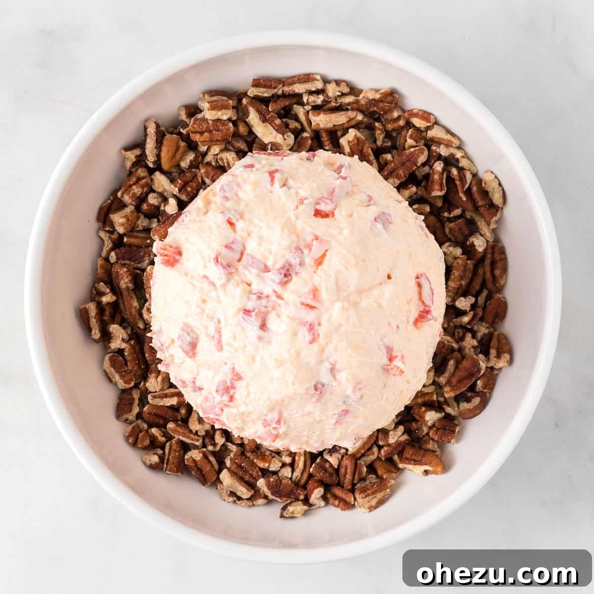 pimento cheese ball being rolled in a bowl filled with chopped pecan pieces for coating