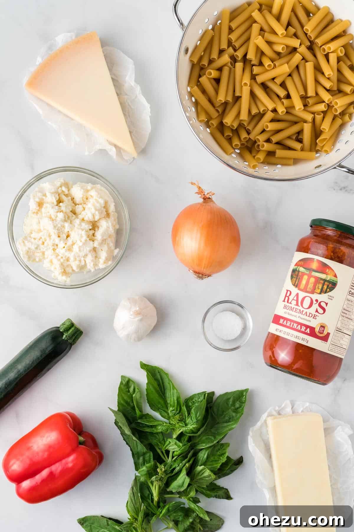 A collection of fresh ingredients laid out, including ziti pasta, yellow onion, garlic, zucchini, red bell pepper, fresh basil, ricotta cheese, parmesan cheese, mozzarella cheese, and a jar of pasta sauce, ready to make vegetarian baked ziti.