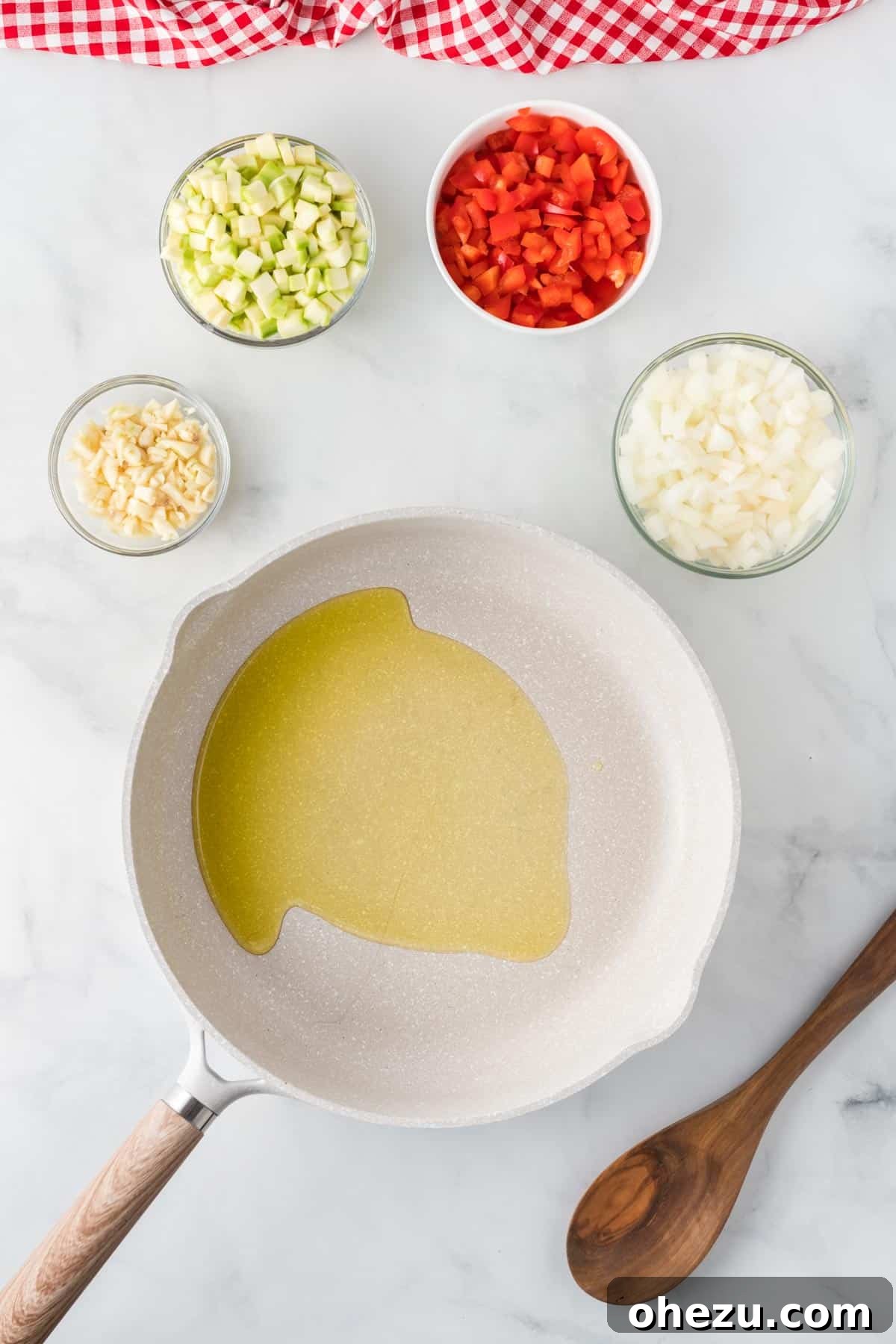 Olive oil heating in a large skillet, surrounded by neatly chopped yellow onion, garlic, zucchini, and red bell pepper, ready for sautéing.