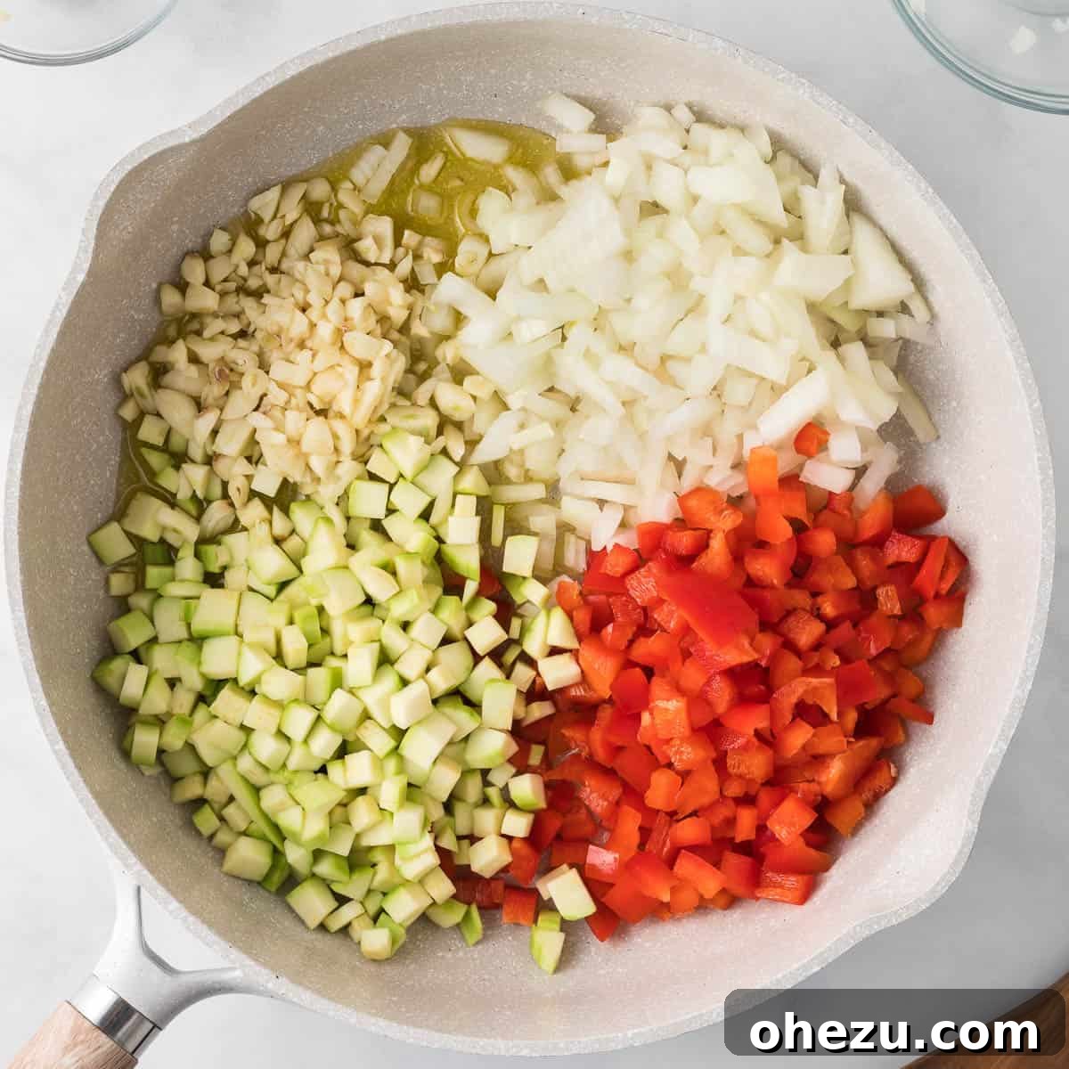 Chopped red bell pepper, onion, garlic, and zucchini being added to a hot skillet with olive oil, ready to be sautéed for the baked ziti.