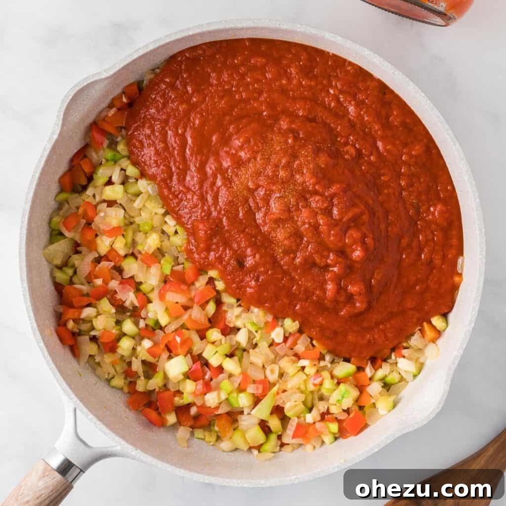 Pouring rich pasta sauce over the sautéed vegetables in a skillet, creating the base for the baked ziti.