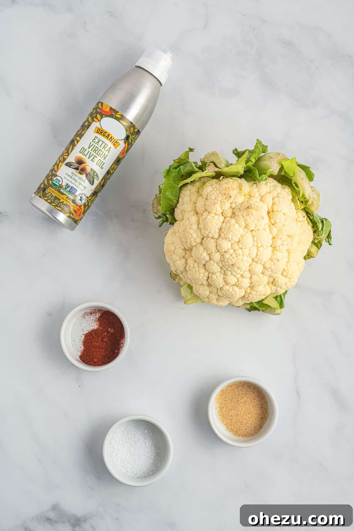 A whole head of fresh cauliflower next to a bottle of olive oil spray and small bowls of salt, garlic powder, and paprika.