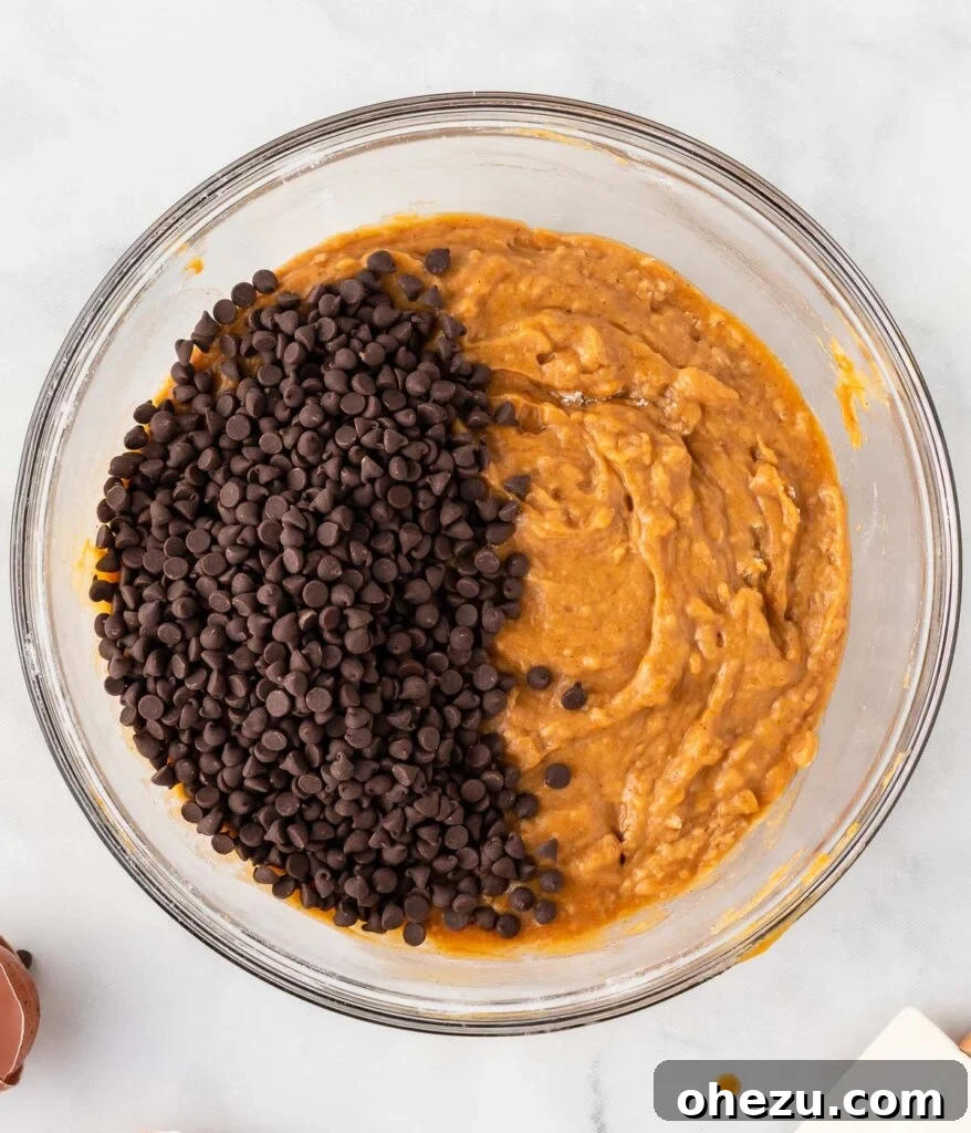 Mini chocolate chips are being gently folded into the rich pumpkin muffin batter using a rubber spatula.