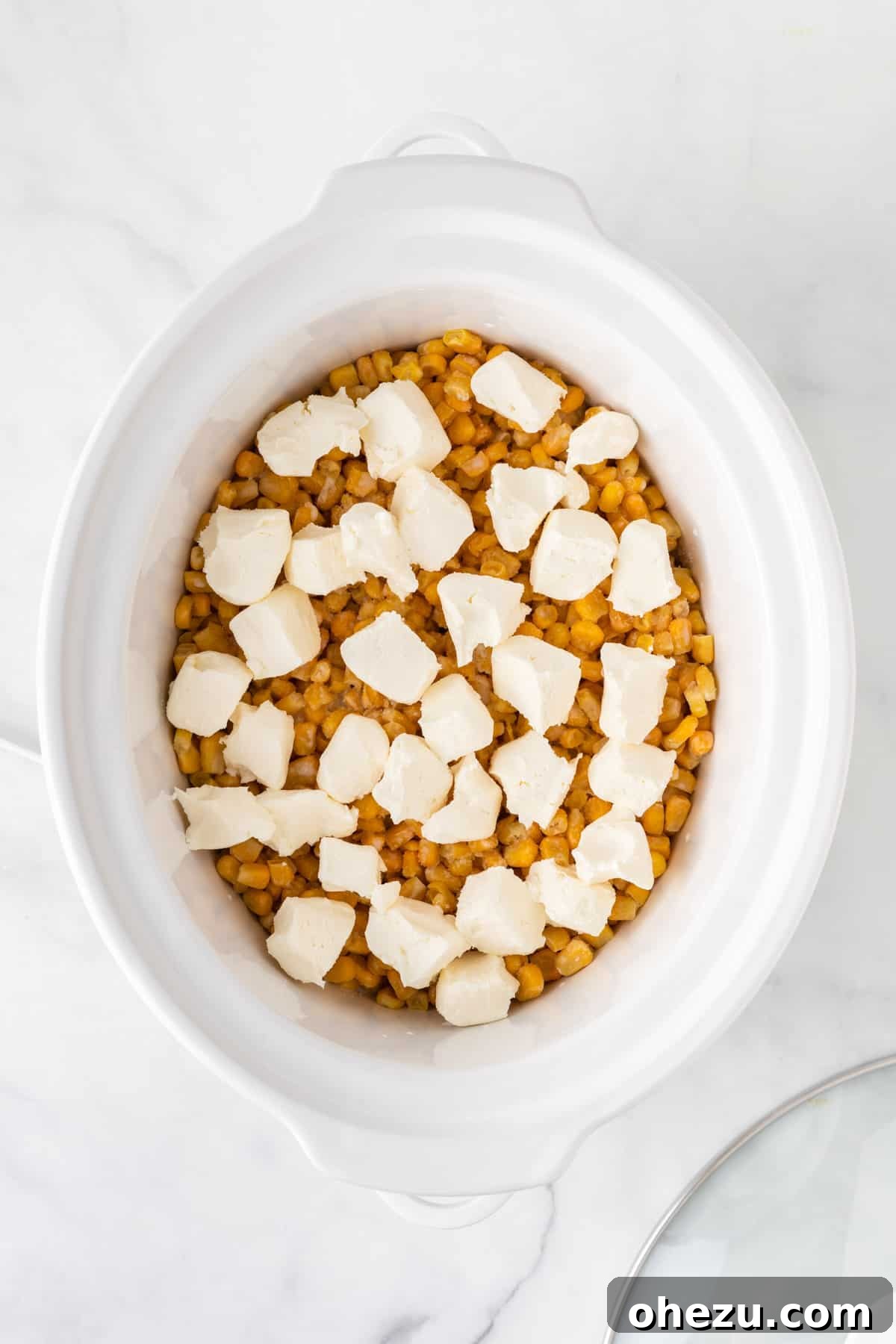 Chunks of cream cheese being placed on top of frozen corn in a slow cooker.