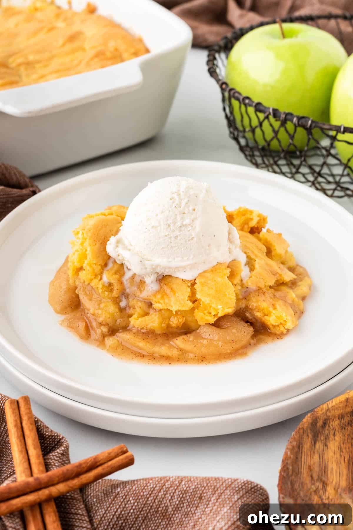 Warm apple cobbler on a white plate, generously topped with a scoop of melting vanilla ice cream and a hint of caramel drizzle.