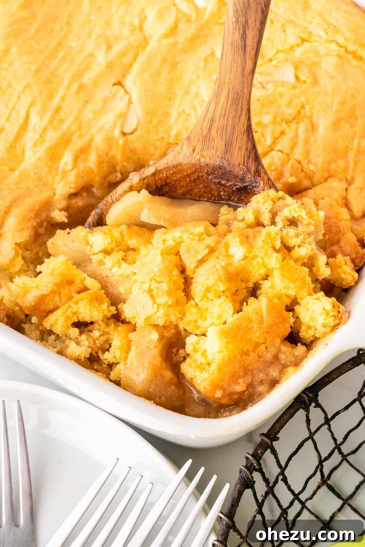 A wooden spoon carefully scooping a generous portion of the warm, bubbly apple cobbler from the baking dish, highlighting the soft cake topping and spiced apple filling.