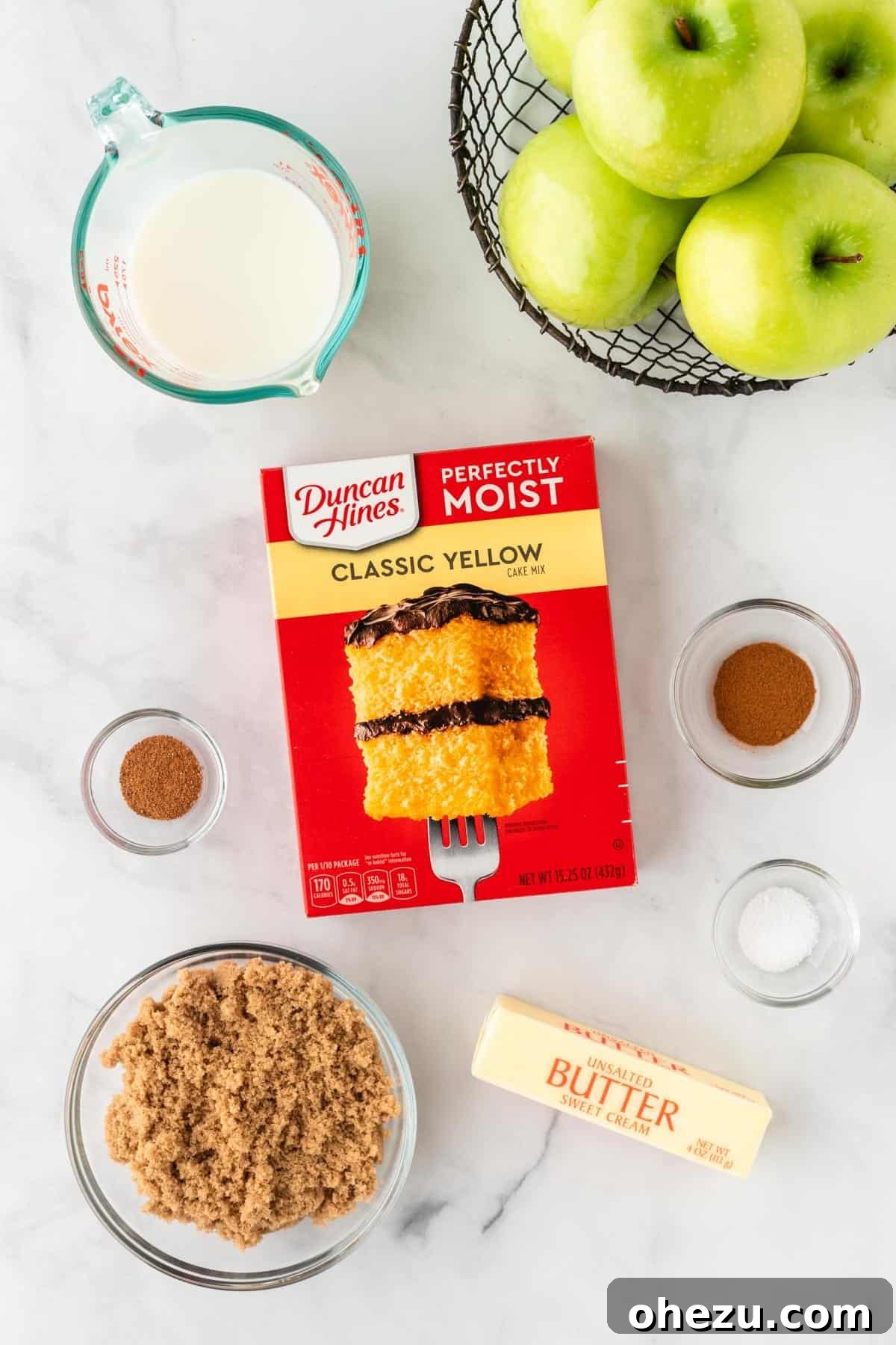 A flat lay photograph showcasing all the fresh ingredients for cake mix apple cobbler: crisp Granny Smith apples, a box of yellow cake mix, brown sugar, butter, milk, cinnamon, nutmeg, and salt.