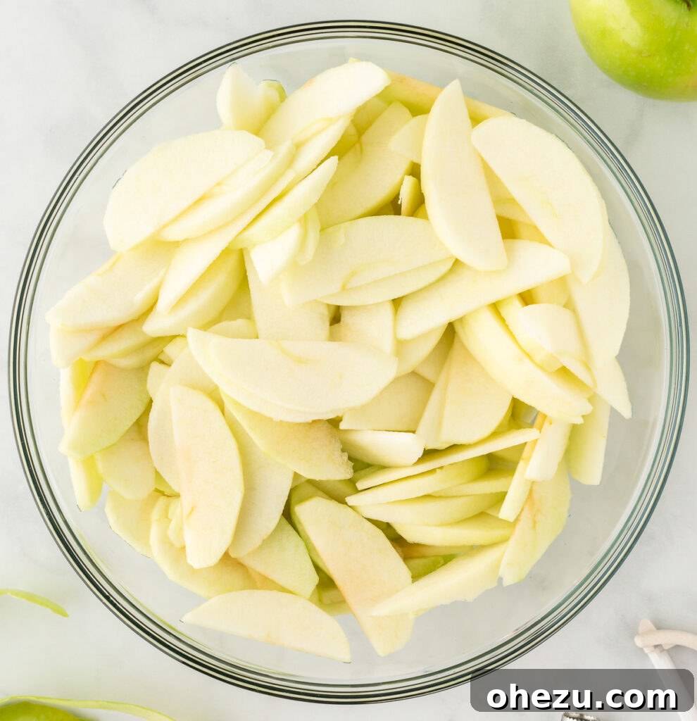 Freshly peeled and thinly sliced Granny Smith apples in a glass mixing bowl, ready for seasoning.