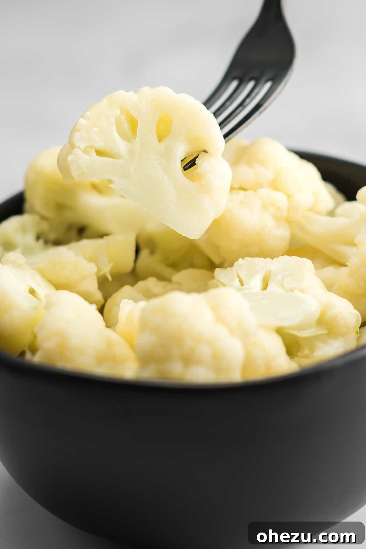A fork picking up a single floret of steamed cauliflower from a bowl.