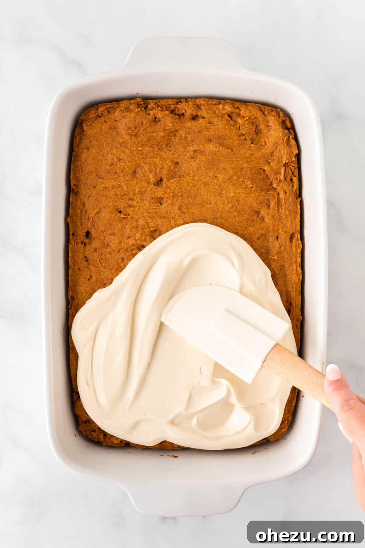 Effortless Fall Spiced Pumpkin Bars 9 An overhead view of a 9x13 inch pan of baked pumpkin bars being frosted with a spatula, showing an even layer of creamy white frosting.
