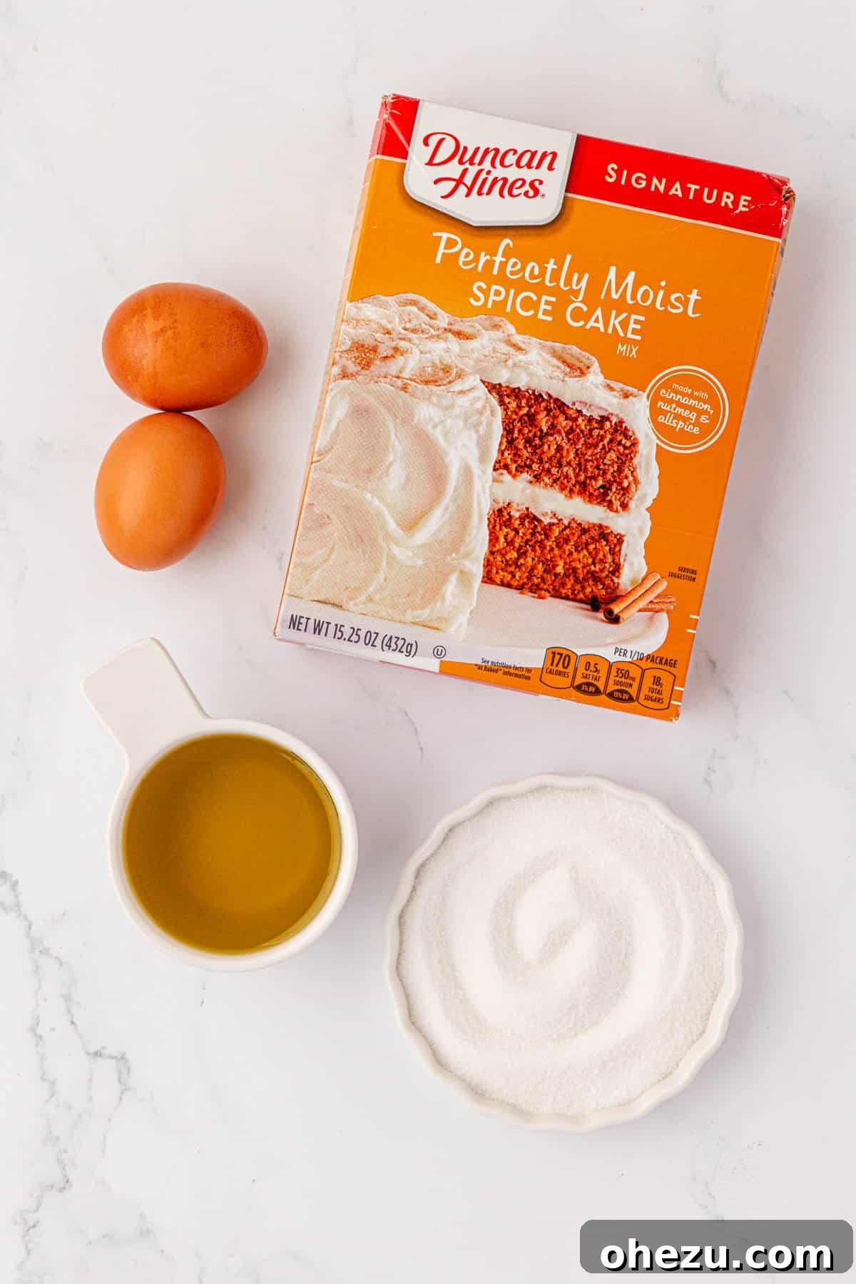 The four essential ingredients for cake mix spice cookies: a box of spice cake mix, fresh eggs, a bottle of olive oil, and a bowl filled with coarse granulated sugar, laid out on a kitchen surface.