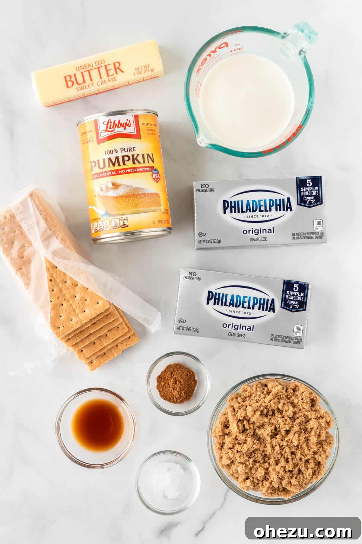ingredients needed to make pumpkin cheesecake bars laid out on a surface, including canned pumpkin, cream cheese, graham crackers, butter, sugar, and spices