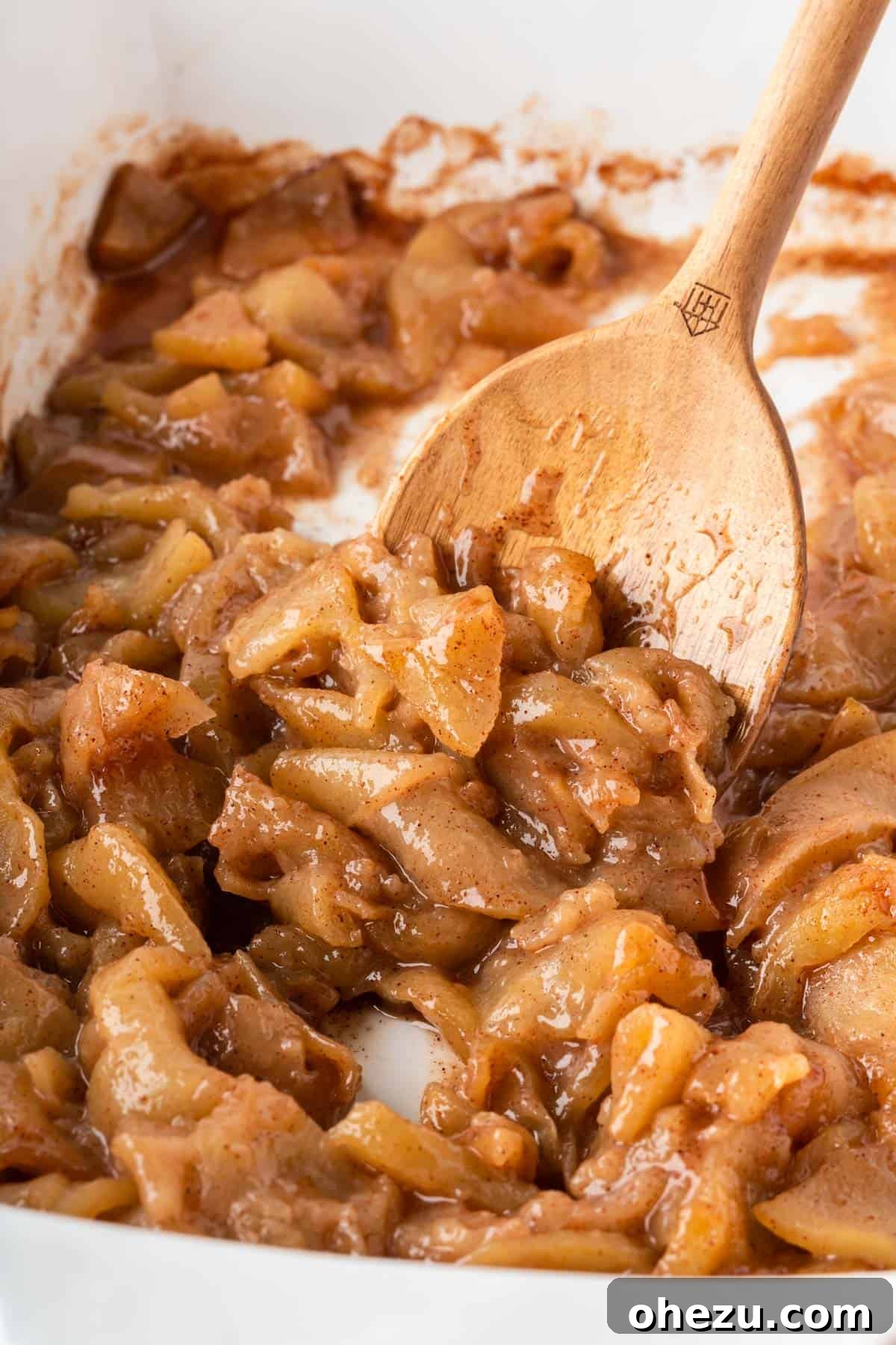 A wooden spoon gently scooping a portion of glistening baked cinnamon apples from the baking dish, highlighting the tender texture and rich sauce.